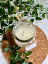 Load image into Gallery viewer, Candle in a glass jar with a label, surrounded by green leaves on a cork board.
